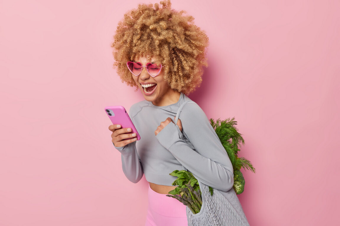 Joyful woman with curly bushy hair uses mobile phone for making shopping online carries fabric bag full of healthy green vegetables for making vegetarian salad isolated over pink background.